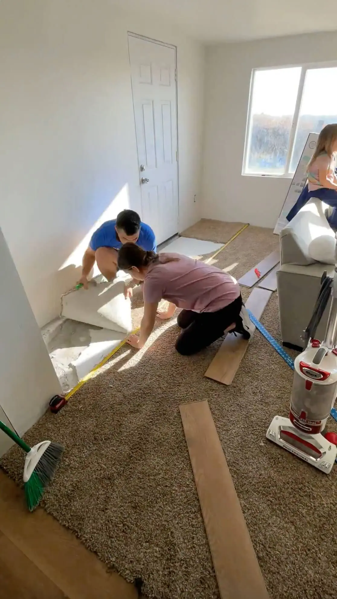 A couple working together to remove old carpet near a doorway, preparing the floor for a new wood-look flooring installation. Tools, planks, and a vacuum are scattered around, showing an active DIY home renovation project.